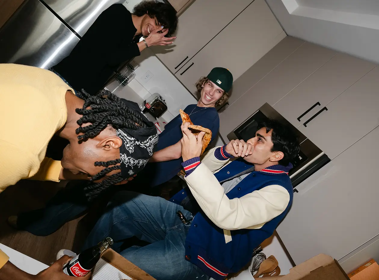 a group of young men laugh while eating pizza inside an apartment kitchen