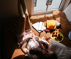 a woman relaxes with food and schoolwork on the floor in a dark room by a window