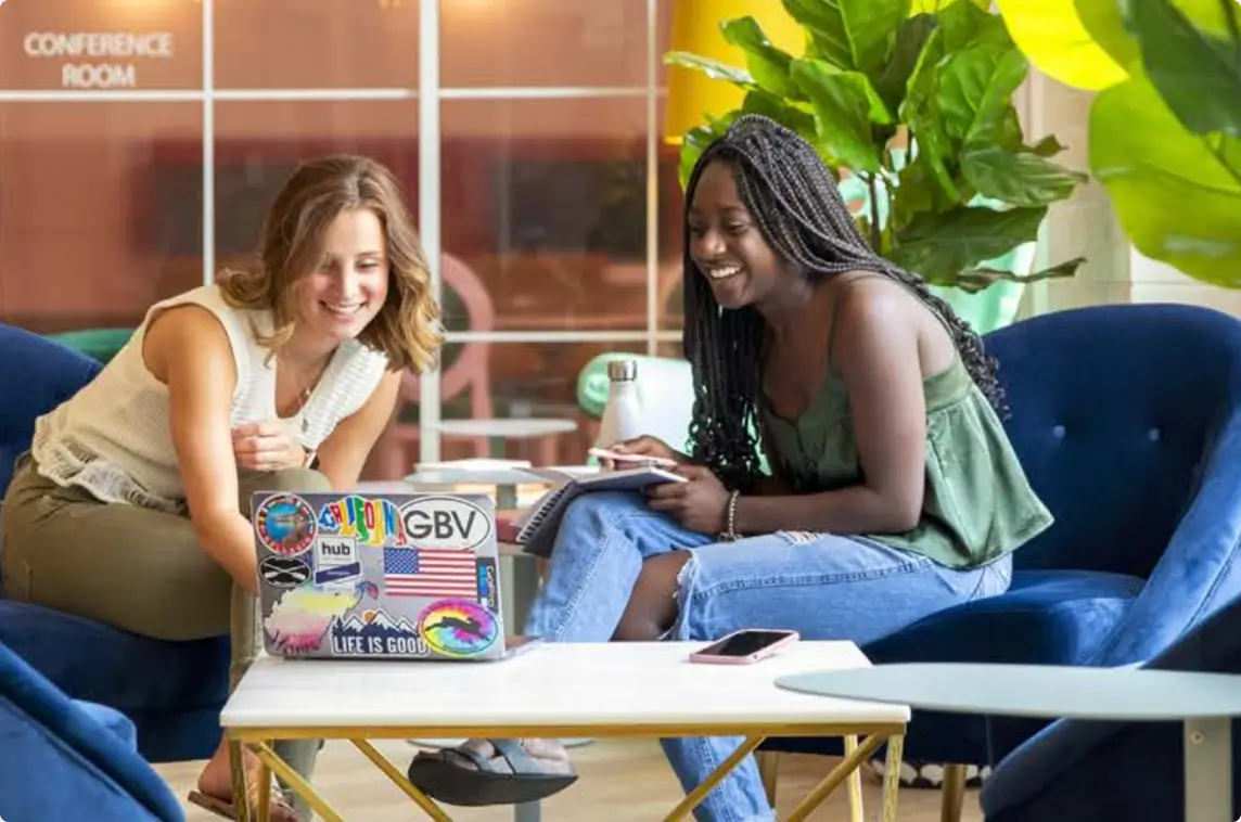 Two women sitting, laughing, and looking at a laptop.